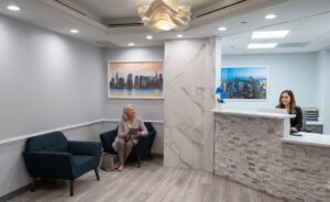 A modern reception area with a woman sitting on a couch and a receptionist at the front desk. The walls are light-colored, with artwork depicting a city skyline.